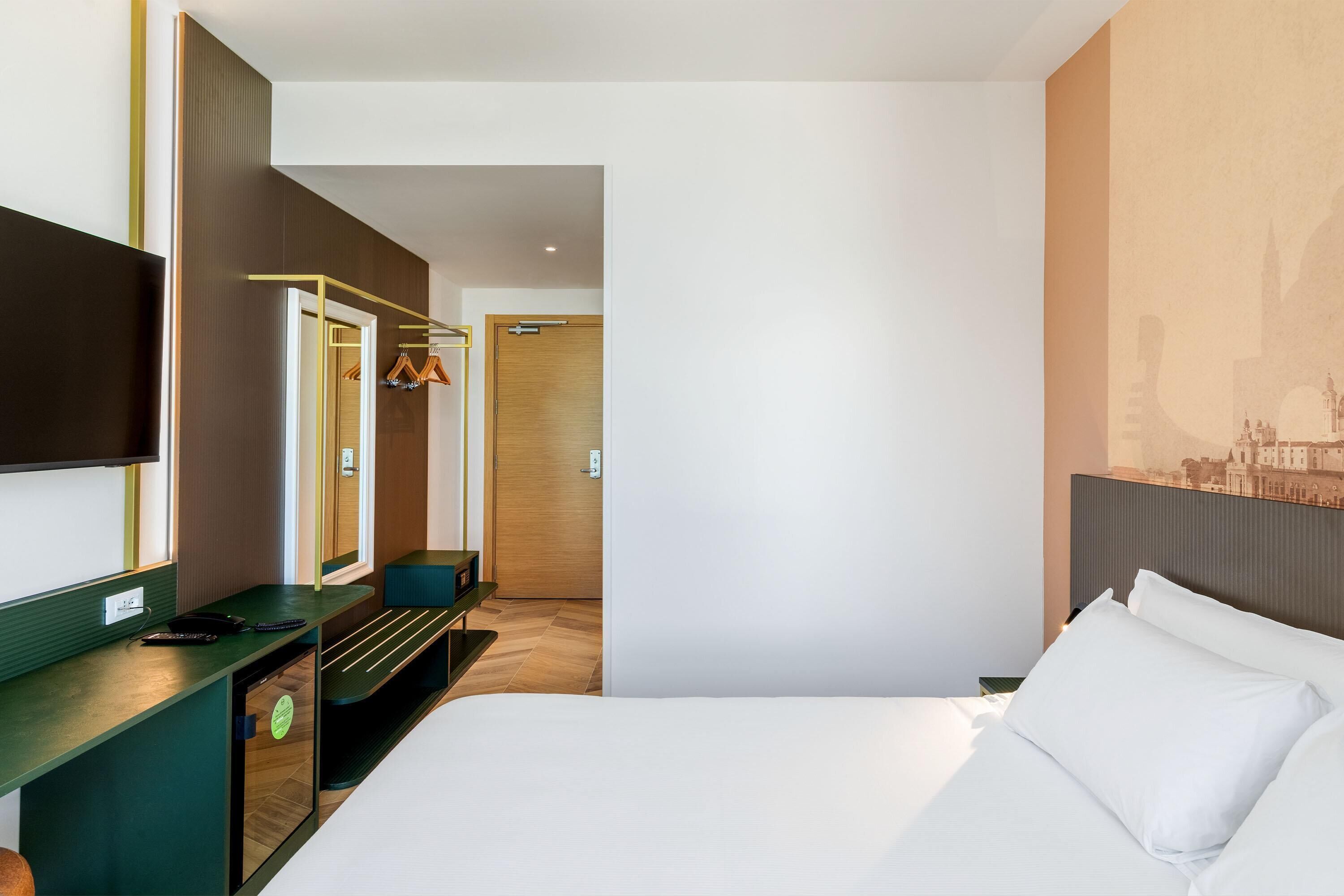 Habitación de hotel con cama, escritorio, televisor, armario y un mural con vistas a la ciudad.