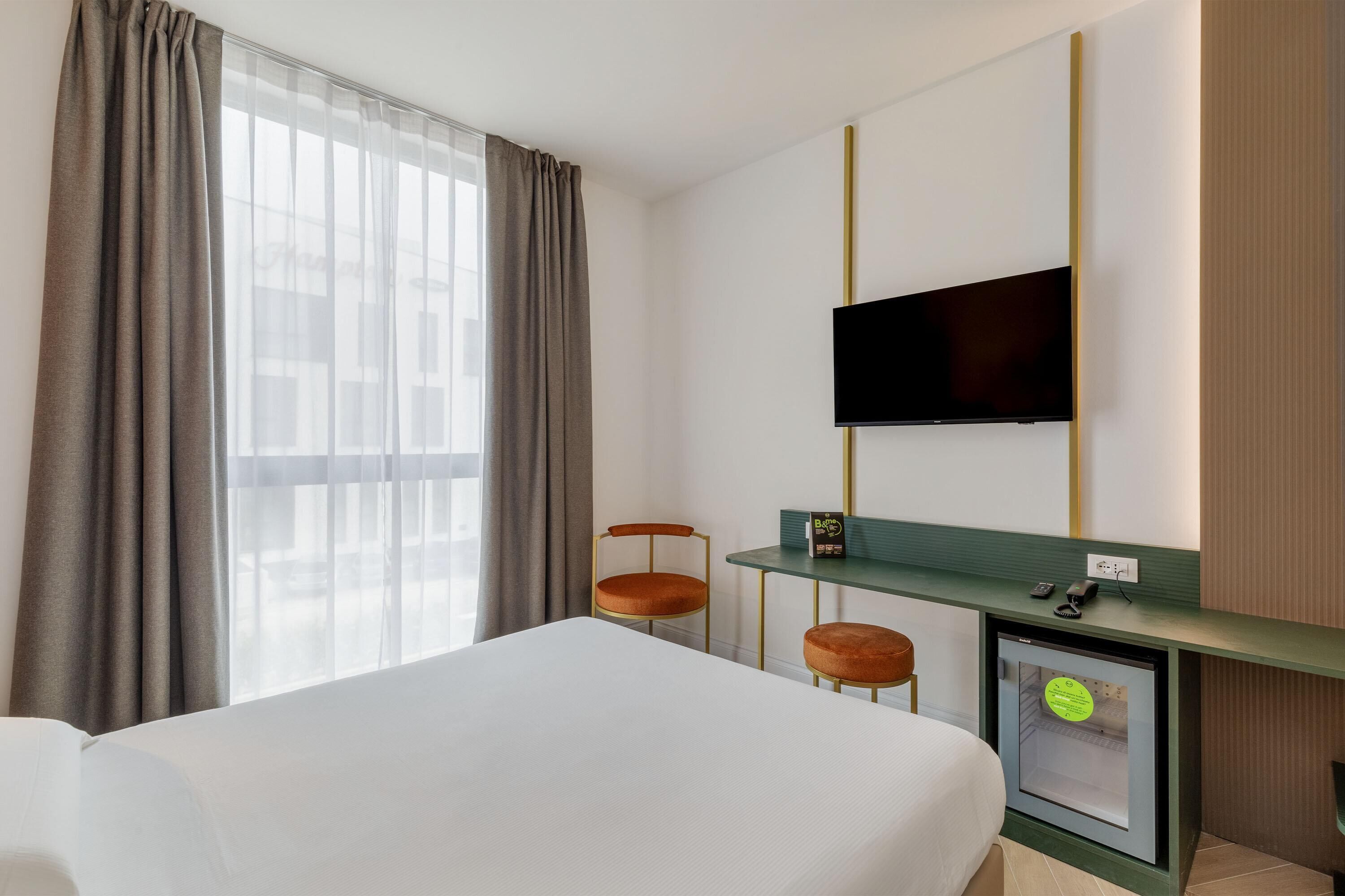 A hotel room with a bed, a TV mounted on the wall, a desk with a chair, and a small cabinet.