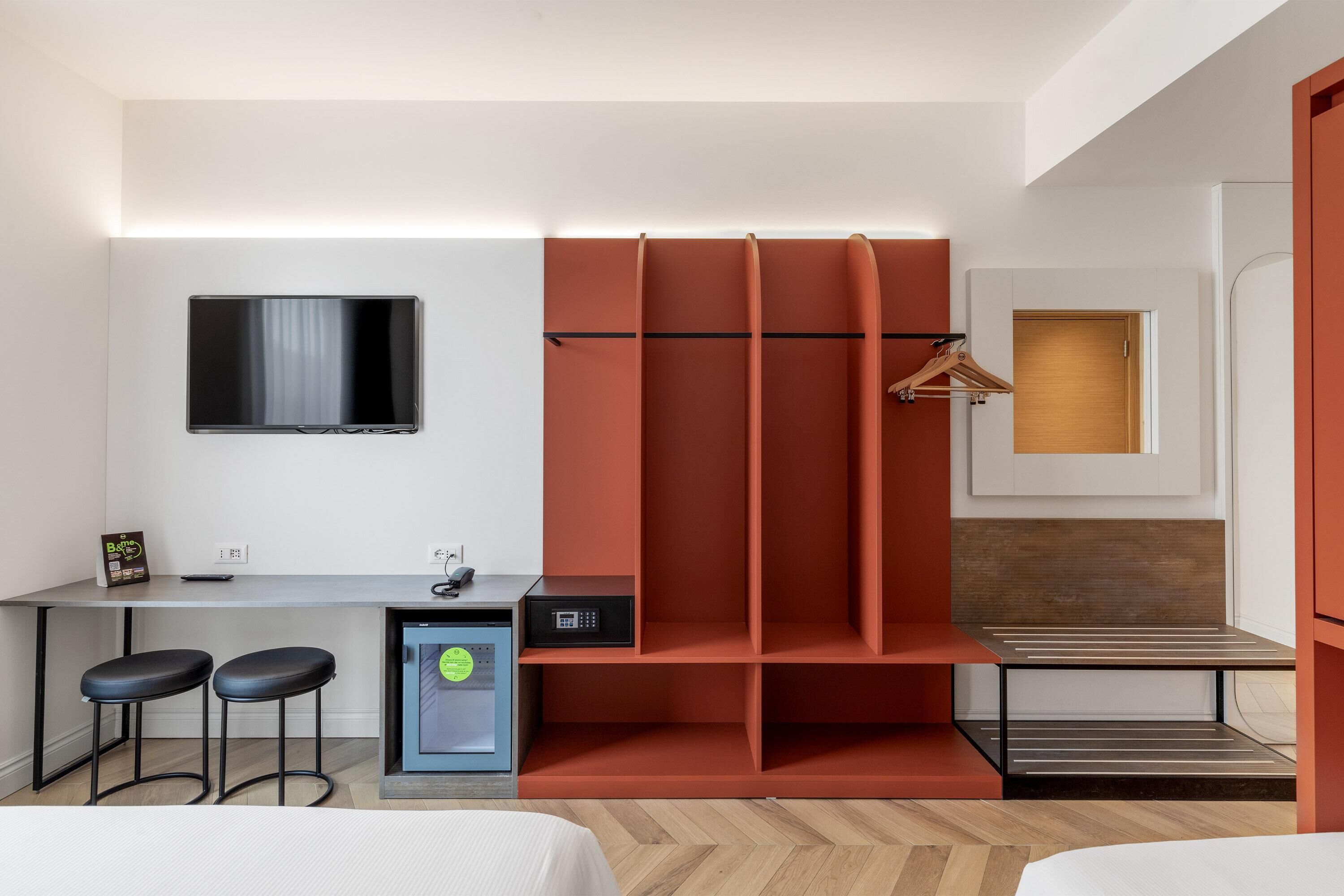 Moderni hotellihuone, jossa on työpöytä, kaksi baarijakkaraa, televisio, minijääkaappi ja peilillä varustettu vaatekaappi.