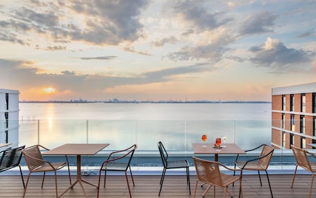 A terrace with tables and chairs overlooking a body of water with a sunset in the background.