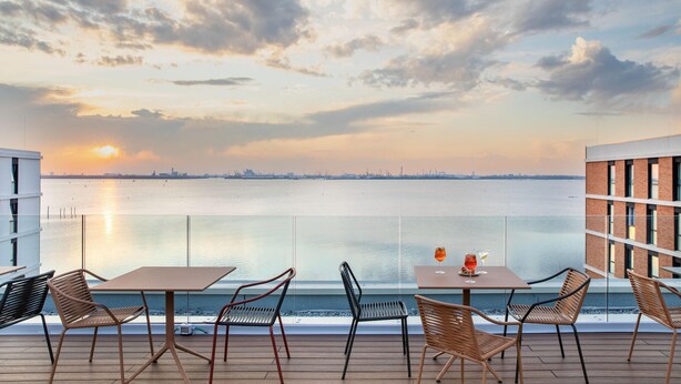 Une terrasse avec des tables et des chaises donnant sur un plan d’eau, avec un coucher de soleil en arrière-plan.