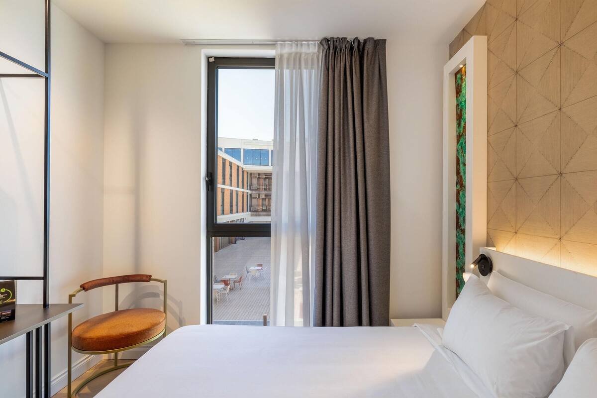 Een hotelkamer met een bed, een stoel, een bureau en uitzicht op een buitenruimte door een raam.