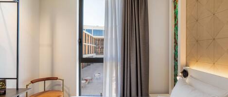 A hotel room with a bed, a chair, a desk, and a view of an outdoor area through a window.