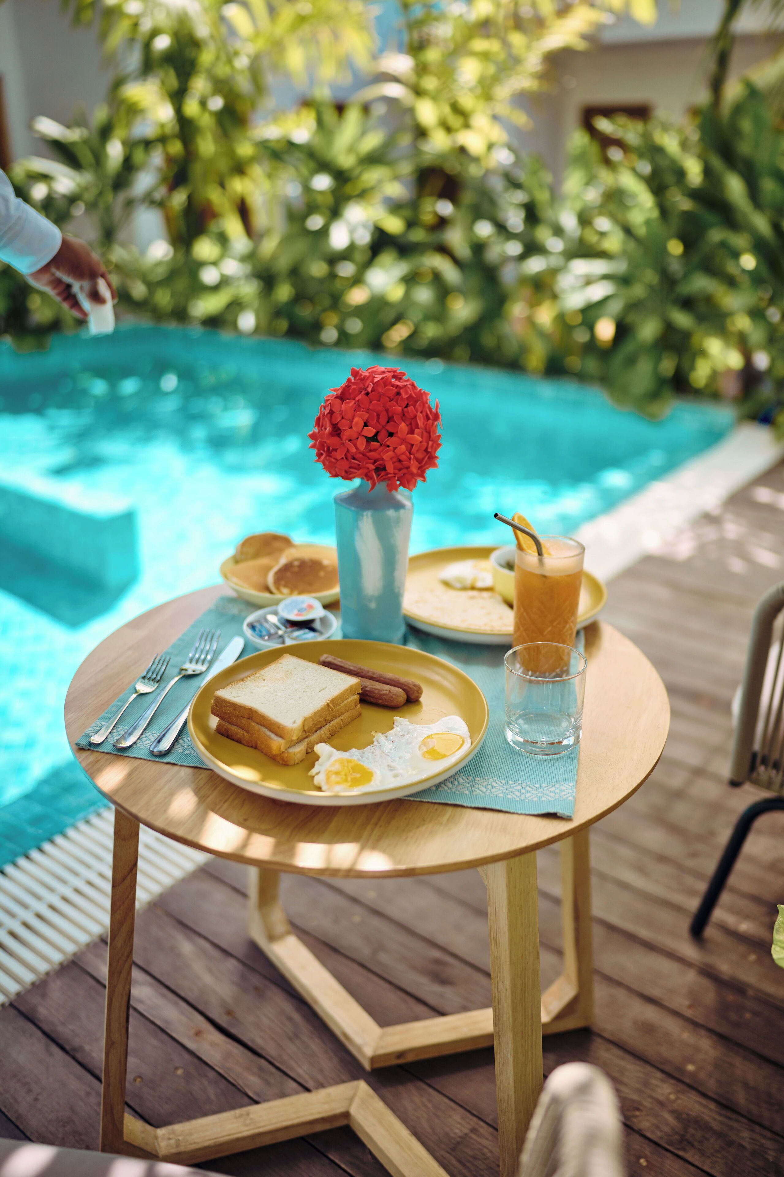 A dining table with breakfast items, a vase with red flowers, and a glass of iced tea by a poolside.