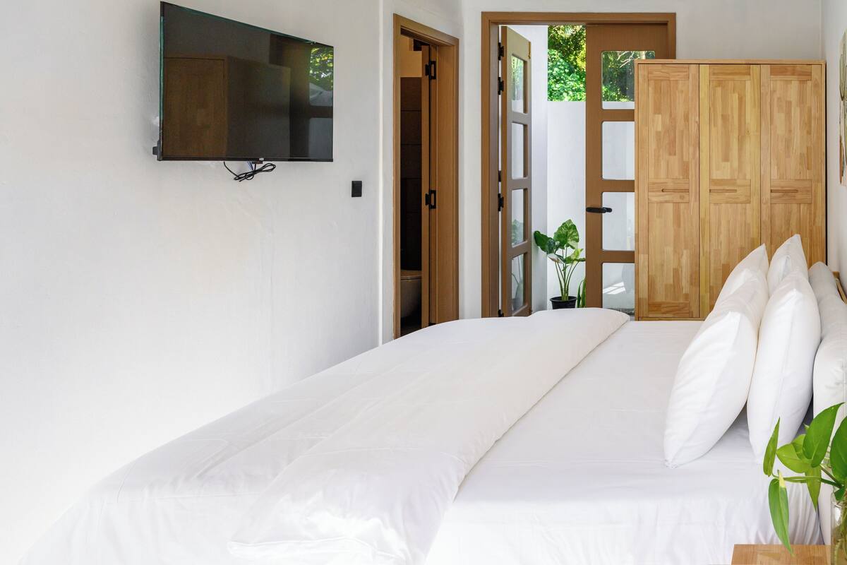 Ein Schlafzimmer mit einem Bett, einem an der Wand befestigten Fernseher, einem Holzschrank und einer Tür zu einem anderen Raum.