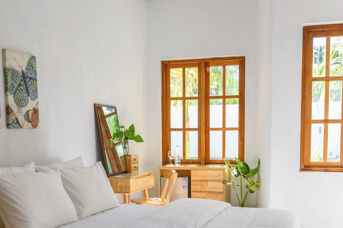 Ein Schlafzimmer mit einem Holzbett, einem Schreibtisch und einem Stuhl. Es gibt Fenster mit Holzrahmen und eine Decke mit sichtbaren Balken.