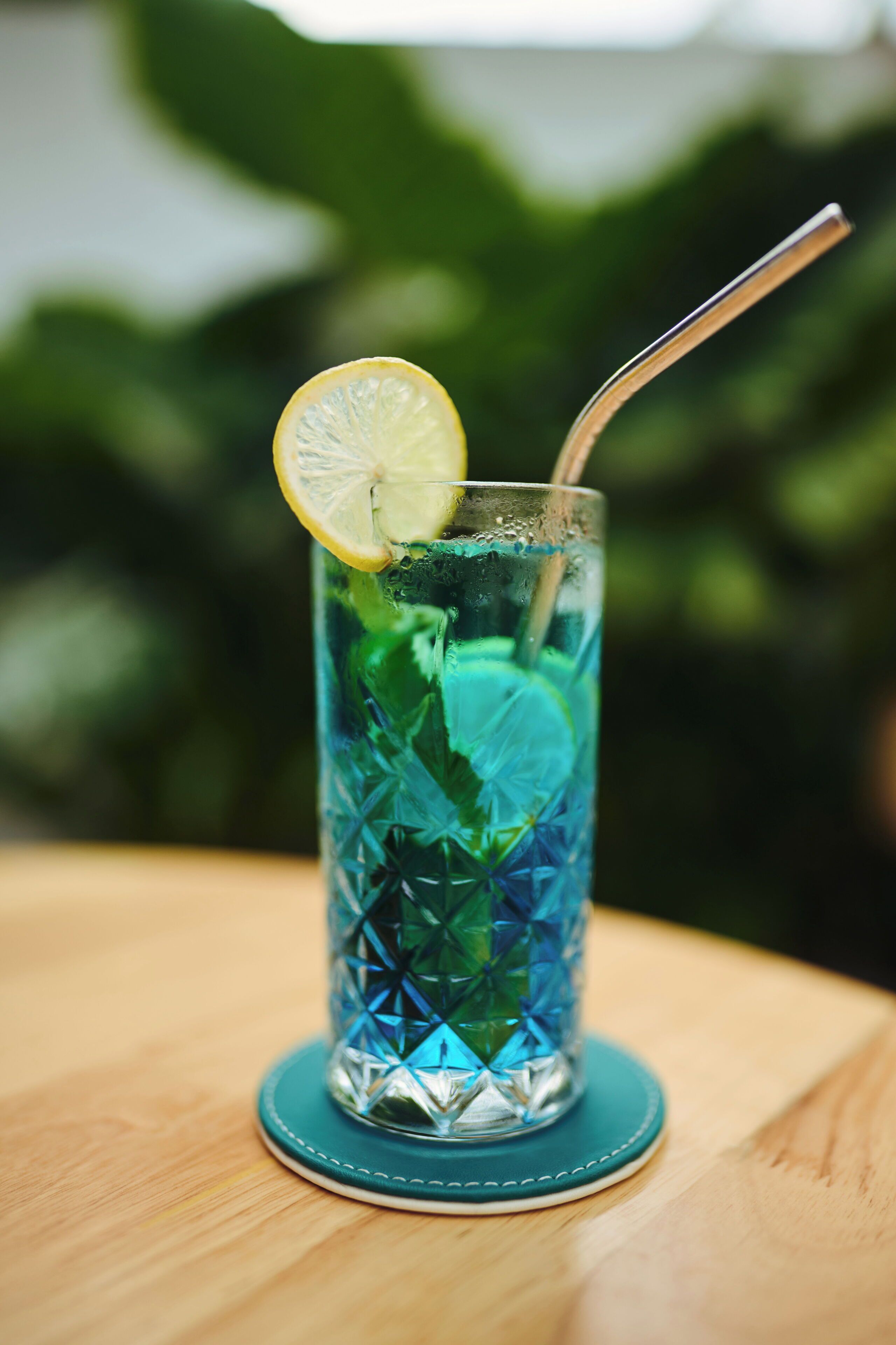 A tall glass with a straw, containing a blue beverage and garnished with a slice of lemon, placed on a wooden table.