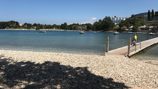 Ein Kiesstrand mit einem hölzernen Steg, der sich über ruhiges Wasser erstreckt, mit Booten und Gebäuden im Hintergrund.
