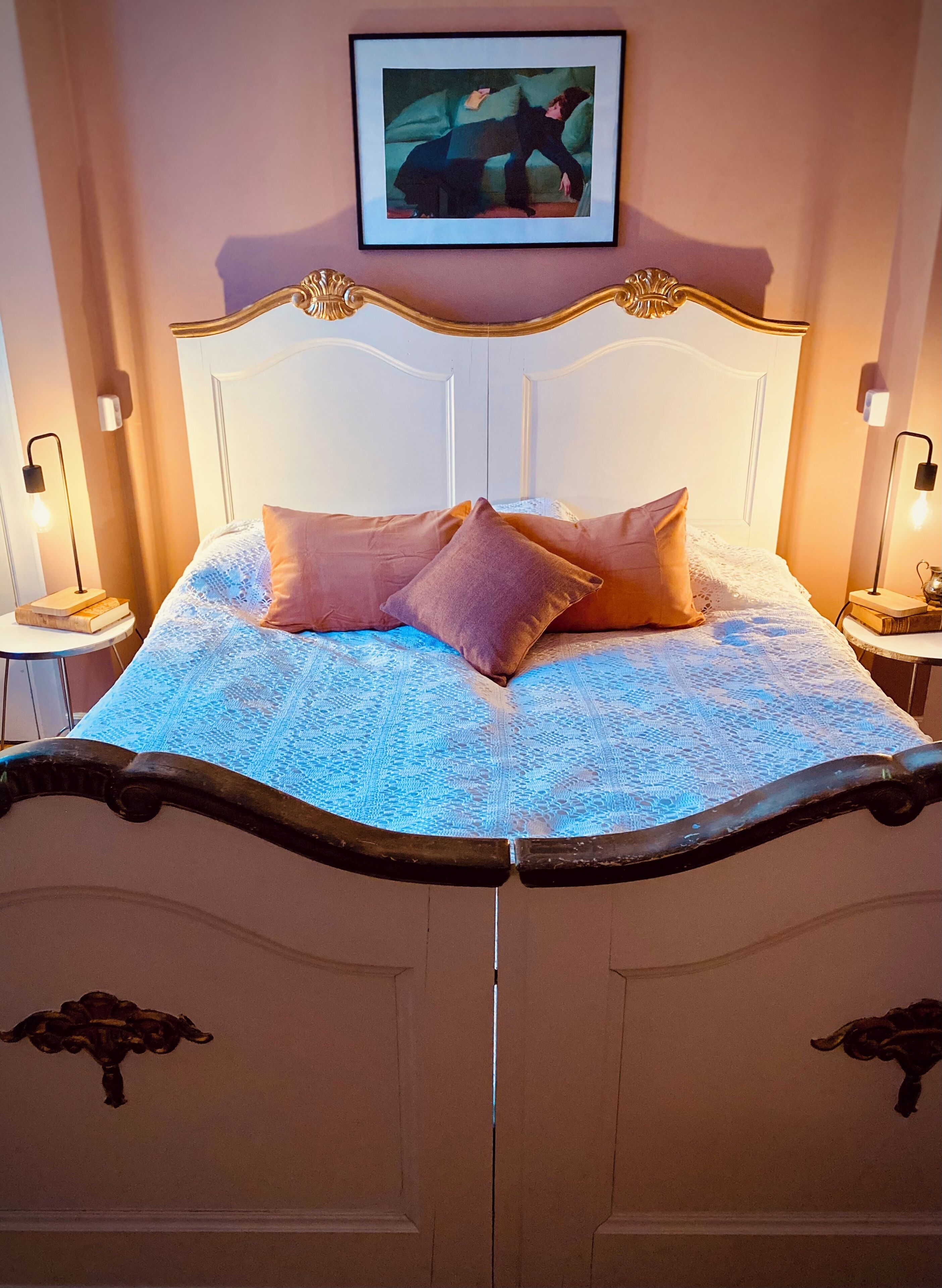 Ein Bett mit weißem Kopfteil, mehreren Kissen und einer blau gemusterten Bettdecke. Es gibt zwei Nachttische mit Lampen und Büchern.