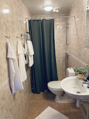 A bathroom with beige tiled walls, a shower with a green curtain, a toilet, and a sink with a plant on the counter.