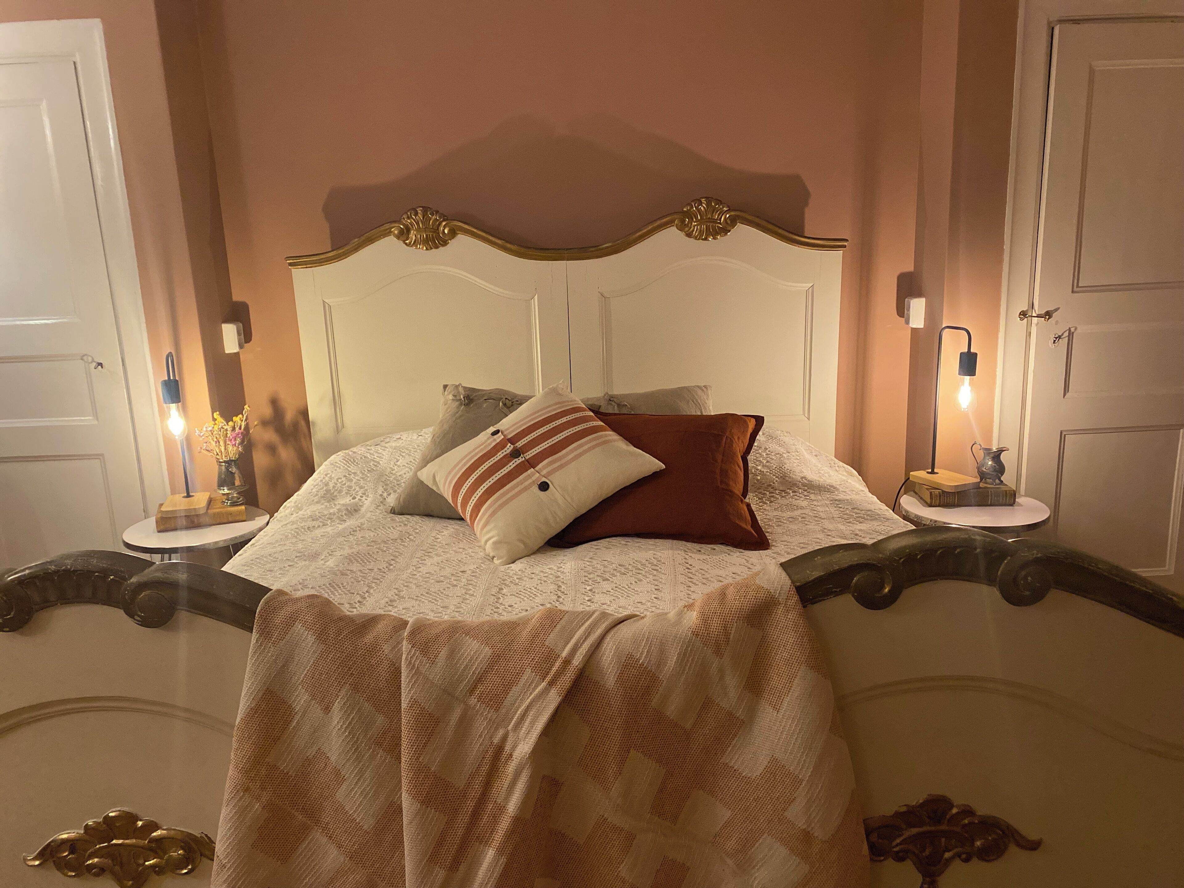 Una cama con cabecera blanca, varios almohadones y una colcha estampada. Hay dos mesitas de noche con lámparas y un pequeño jarrón con flores.