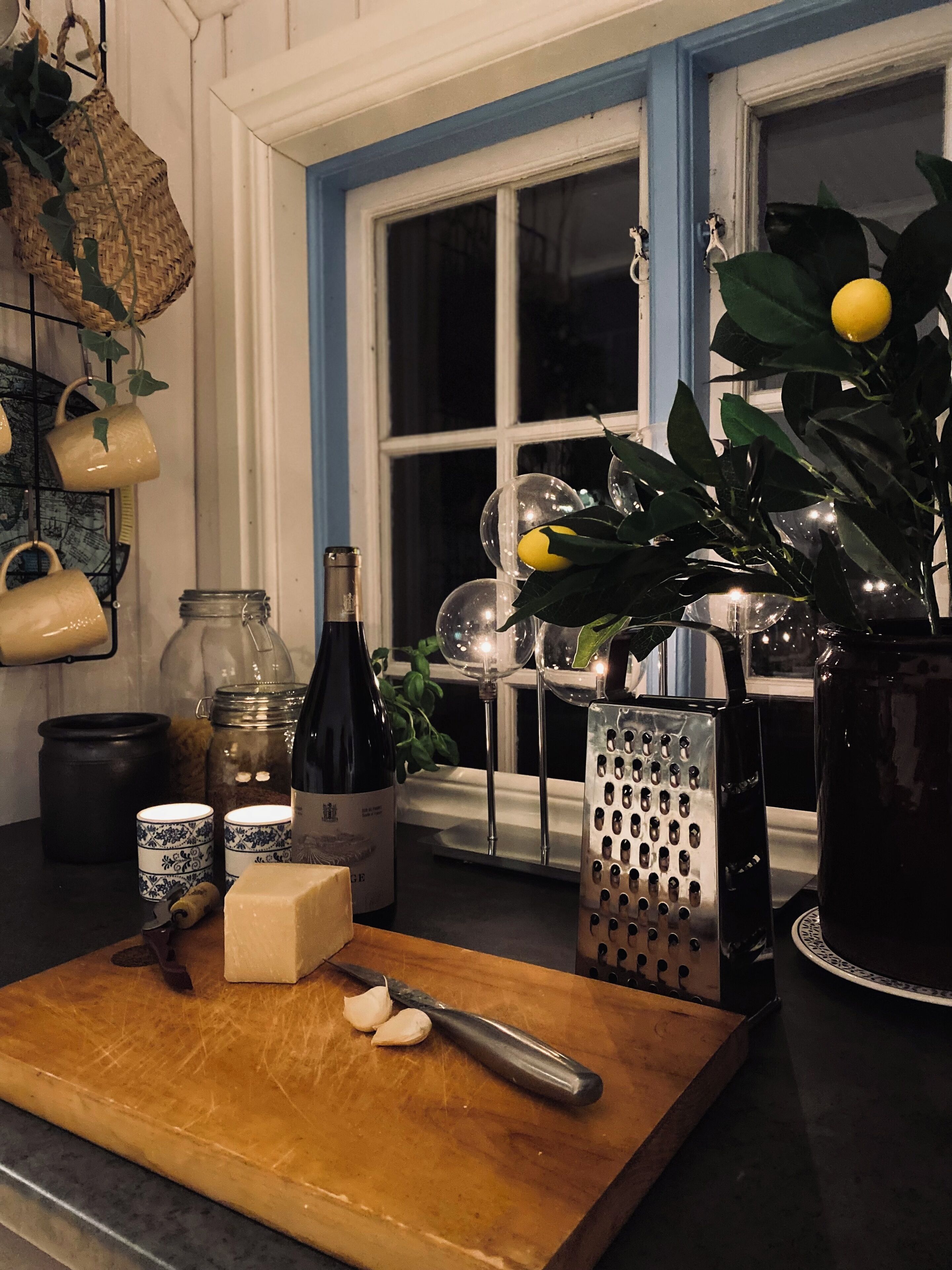 A kitchen counter with a wooden cutting board, a knife, a grater, a bottle of wine, and a vase with lemons.