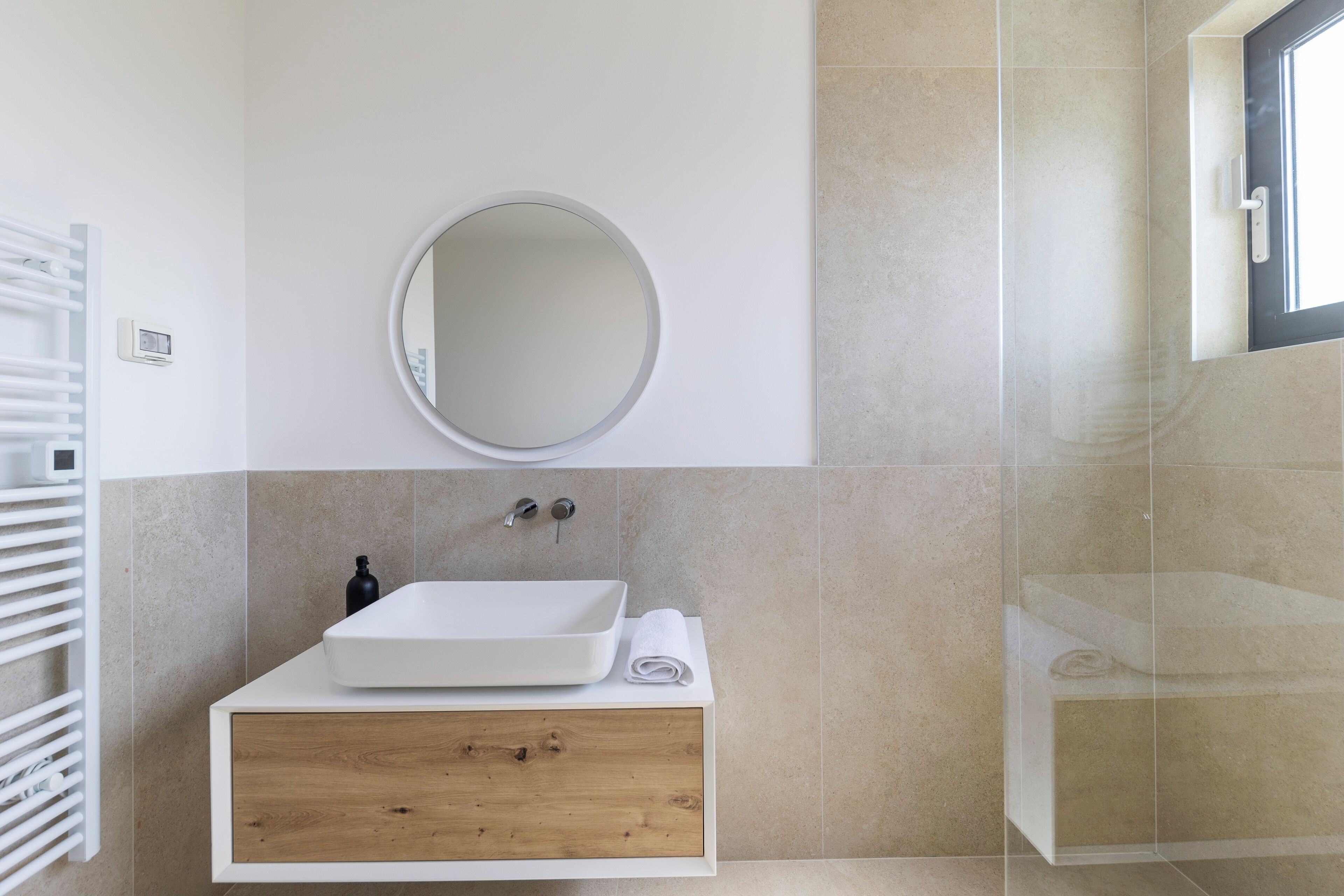A modern bathroom with a white sink, wooden drawer, and a round mirror.
