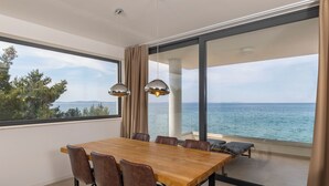 Een moderne eetruimte met een houten tafel en stoelen, grote ramen met uitzicht op zee en hanglampen.