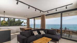 Een moderne woonkamer met een hoekbank, een houten salontafel en een eetgedeelte met rieten stoelen. Grote ramen bieden uitzicht op zee.