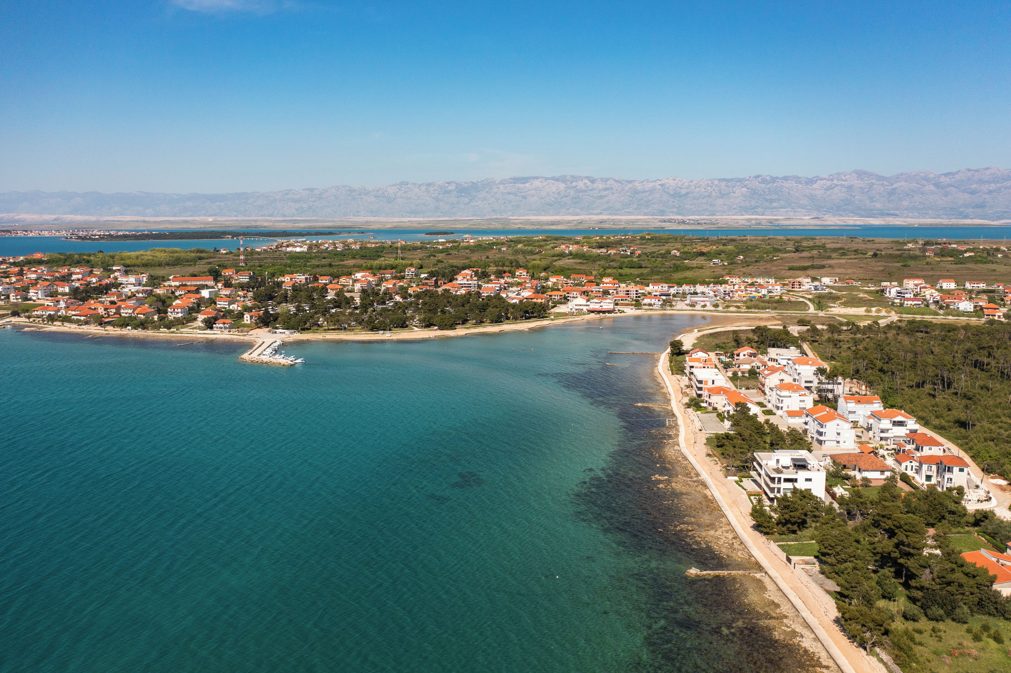 An aerial view of a coastal town with a marina, houses with red roofs, and a clear view of the sea and mountains in the background.
