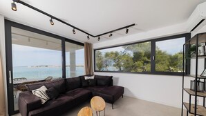 Een moderne woonkamer met een donkere hoekbank, twee houten bijzettafeltjes en grote ramen met uitzicht op zee.