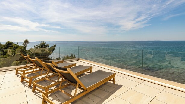 Een terras met houten loungestoelen en een glazen balustrade met uitzicht op een waterpartij.