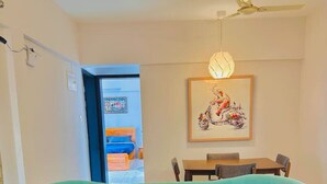A teal sofa with yellow legs in a room with a dining table and chairs, a ceiling fan, and a framed artwork on the wall. - Flamingo: 2BHK AC Flat, Near Phoenix Mall Wakad (Pimpri-Chinchwad)