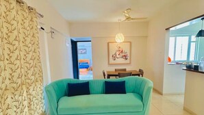 A living room with a teal sofa, a dining table, and a kitchen area. - Flamingo: 2BHK AC Flat, Near Phoenix Mall Wakad (Pimpri-Chinchwad)