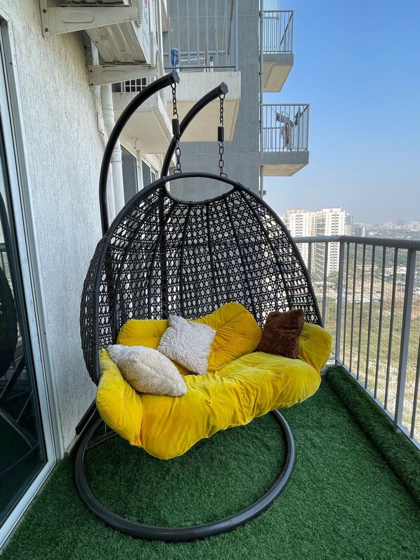 Een hangstoel met gele kussens op een balkon met uitzicht op gebouwen.