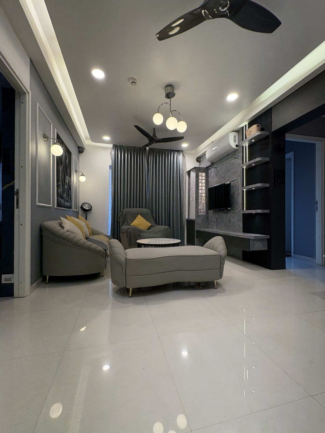 A modern living room with a sofa, armchair, and ottoman, a ceiling fan, recessed lighting, and a flat-screen TV.