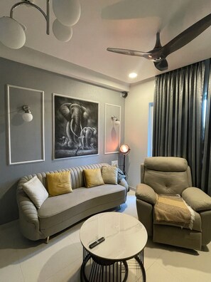 Un salon moderne avec un canapé, un fauteuil et une table basse ronde. Les murs sont ornés de tableaux encadrés, et on y trouve également un ventilateur de plafond et une lampe sur pied.