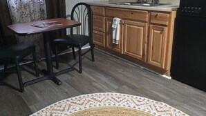 A kitchen with wooden cabinets, a black refrigerator, a dining table with two chairs, and a sign that reads "Welcome to our Neck of the Woods."