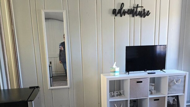A modern living room with a flat-screen TV on a white stand, a mirror, and a wall-mounted sign that reads "adventure".