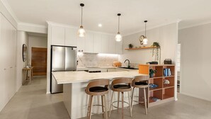 Una cocina moderna con una isla central, taburetes de bar, nevera integrada, luces colgantes y un estante de madera con libros y plantas.