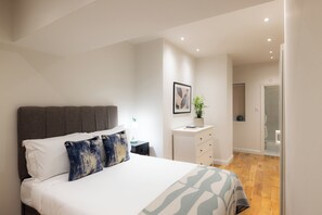 A bedroom with a bed, a bedside table, a dresser, and a bathroom visible through an open door. - The Maddox by Nestor (London)