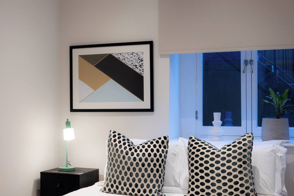 Une chambre à coucher avec un lit, une table de chevet avec une lampe, une œuvre d’art abstraite encadrée et une fenêtre donnant sur un escalier et une plante.