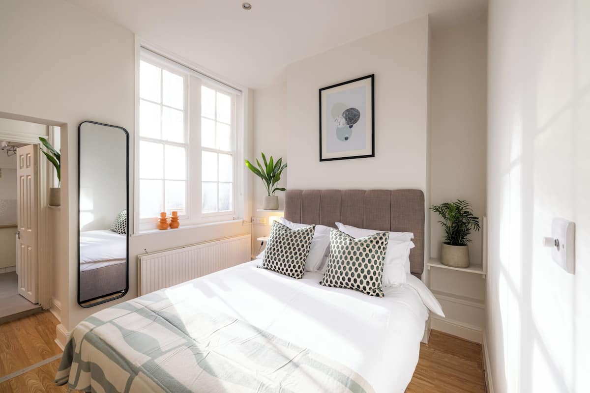 Une chambre à coucher avec un lit, un grand miroir, une fenêtre, une plante en pot et un tableau encadré accroché au mur.