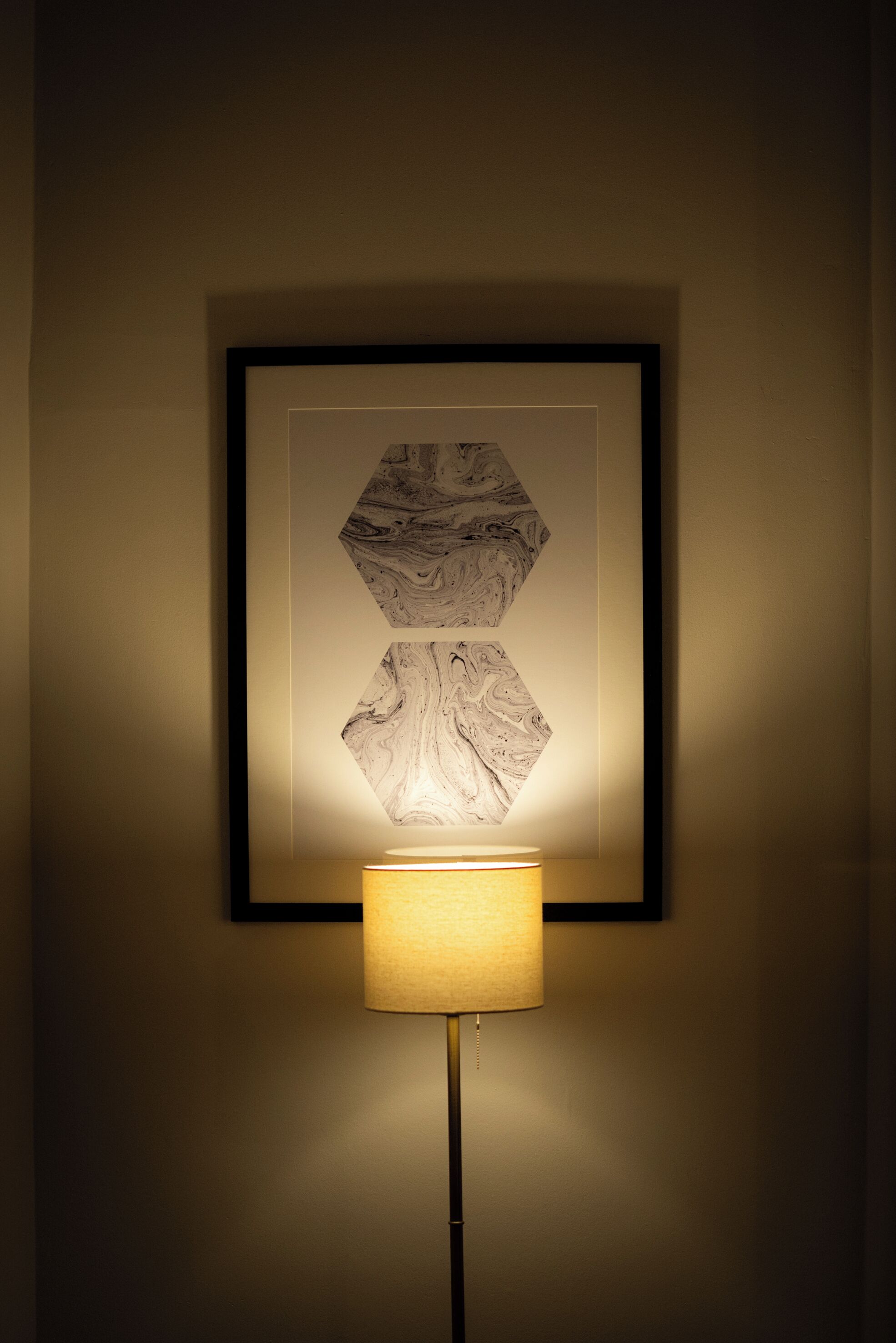 A framed artwork with a geometric pattern on a wall, illuminated by a floor lamp.