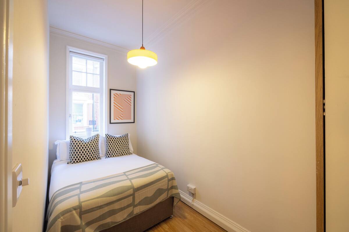 Une chambre à coucher avec un lit, une fenêtre, un luminaire fixé au mur et un tableau encadré.