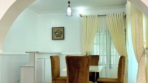 Un comedor con una mesa blanca, cuatro sillas marrones, un mueble blanco pequeño y una lámpara colgante.