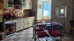 Une cuisine avec un mur recouvert de carreaux, des meubles en bois, une cuisinière et un coin repas comprenant une table en verre et des chaises en métal.