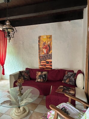 A living room with a red sofa, a glass coffee table, a wooden chair, a wall-mounted painting of a squirrel, and a red curtain. - Typical Cévenole house with breathtaking views (Pied-de-Borne)