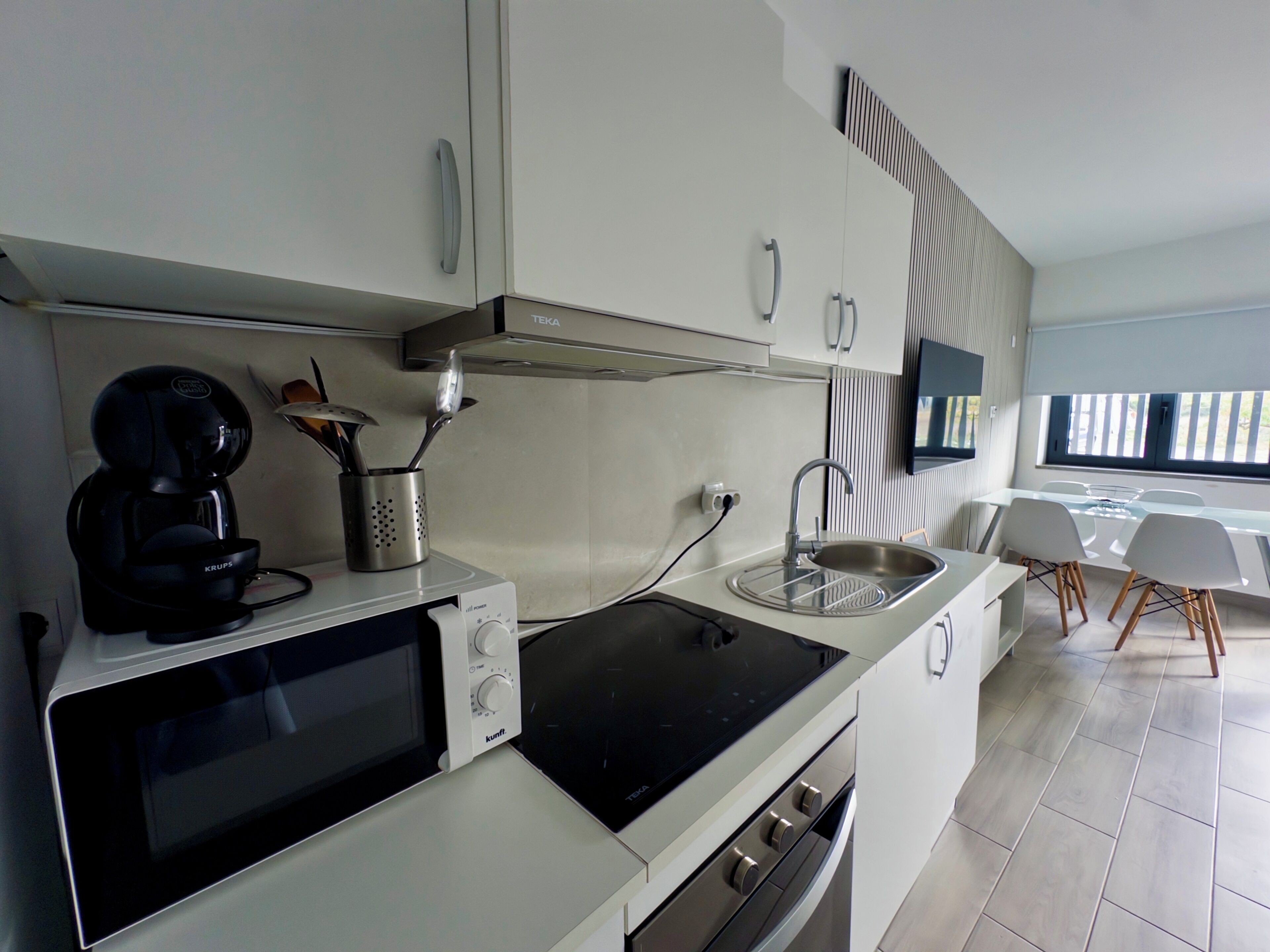 A modern kitchen with a microwave, stove, and sink. There is a coffee maker and utensils on the counter. A TV is mounted on the wall, and a dining area with chairs is visible in the background.