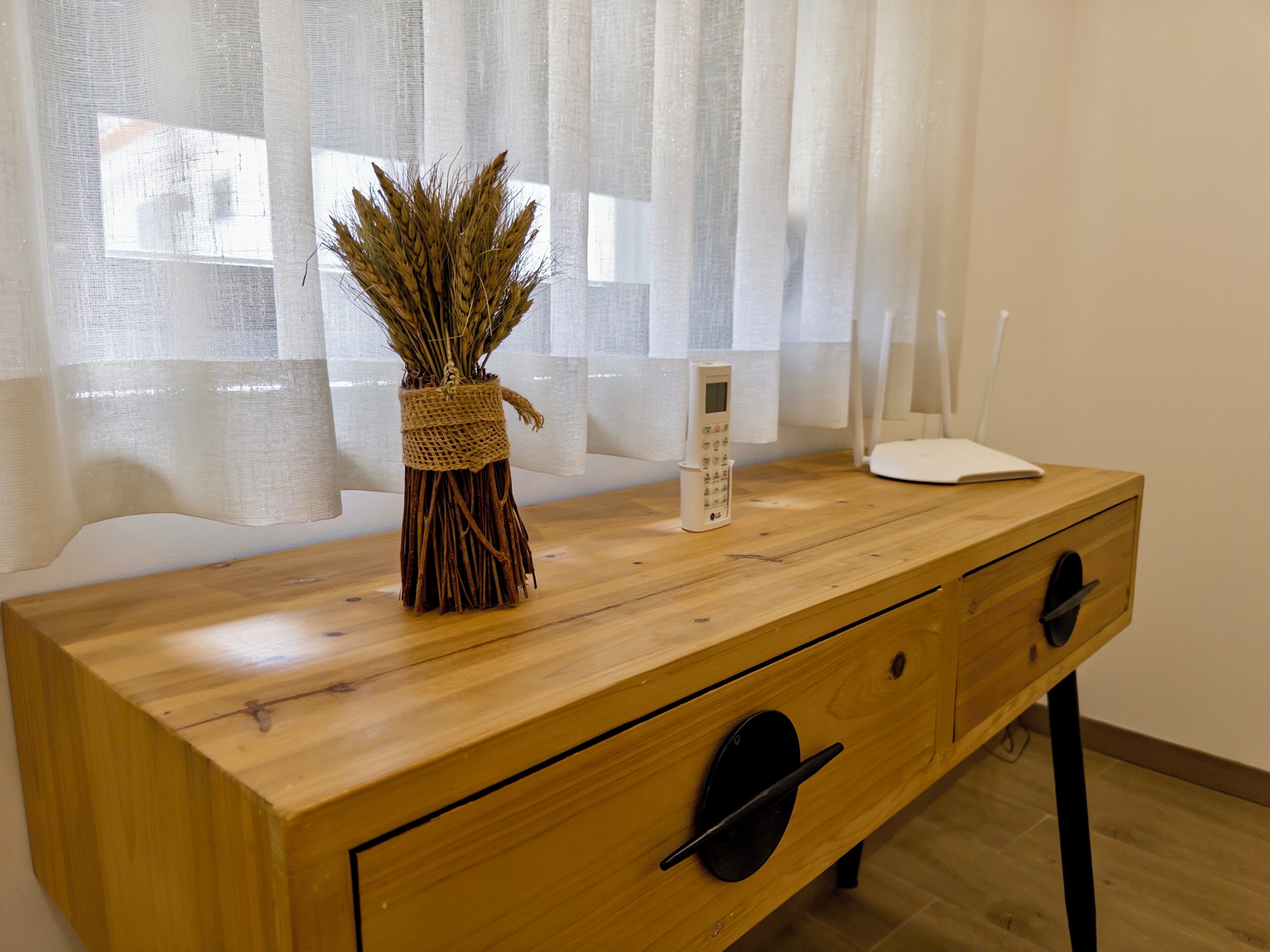 A wooden desk with a vase of dried plants, a remote control, and a white device on it.