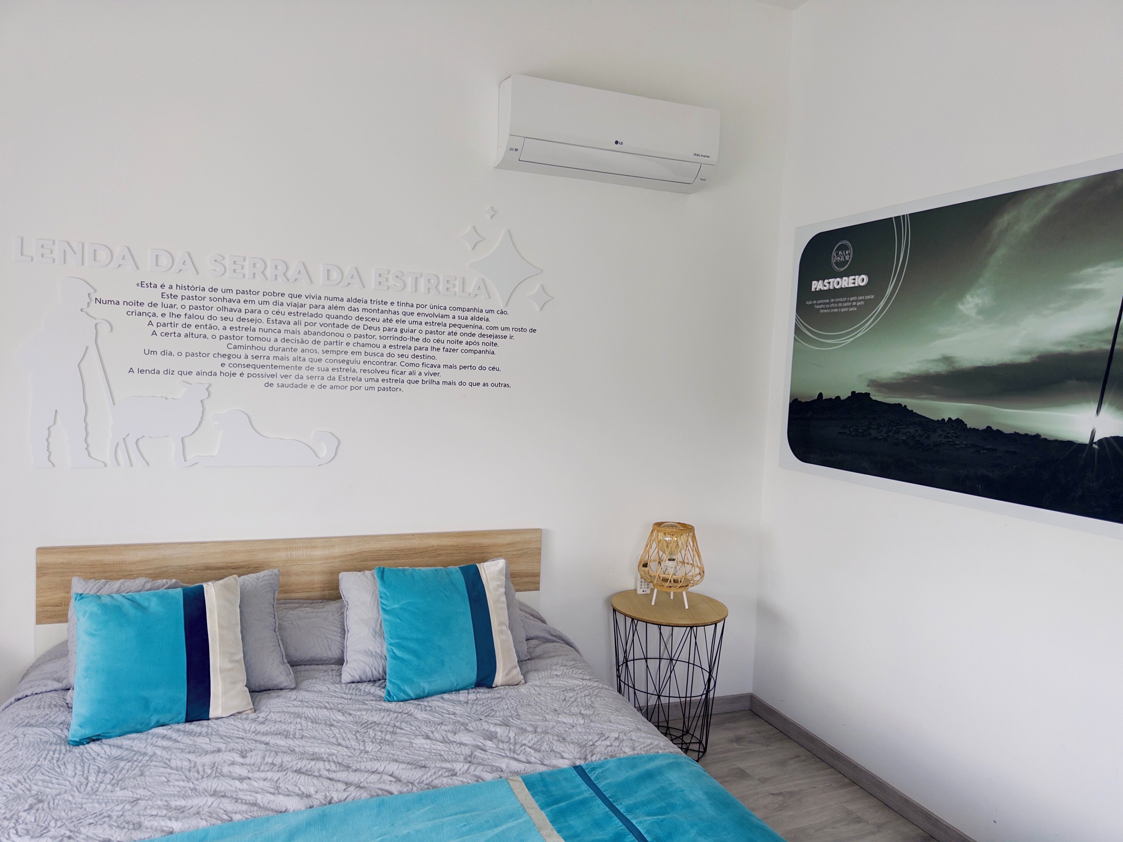 A bedroom with a bed, pillows, a wall-mounted air conditioner, a wall art piece, and a small side table with a lamp.
