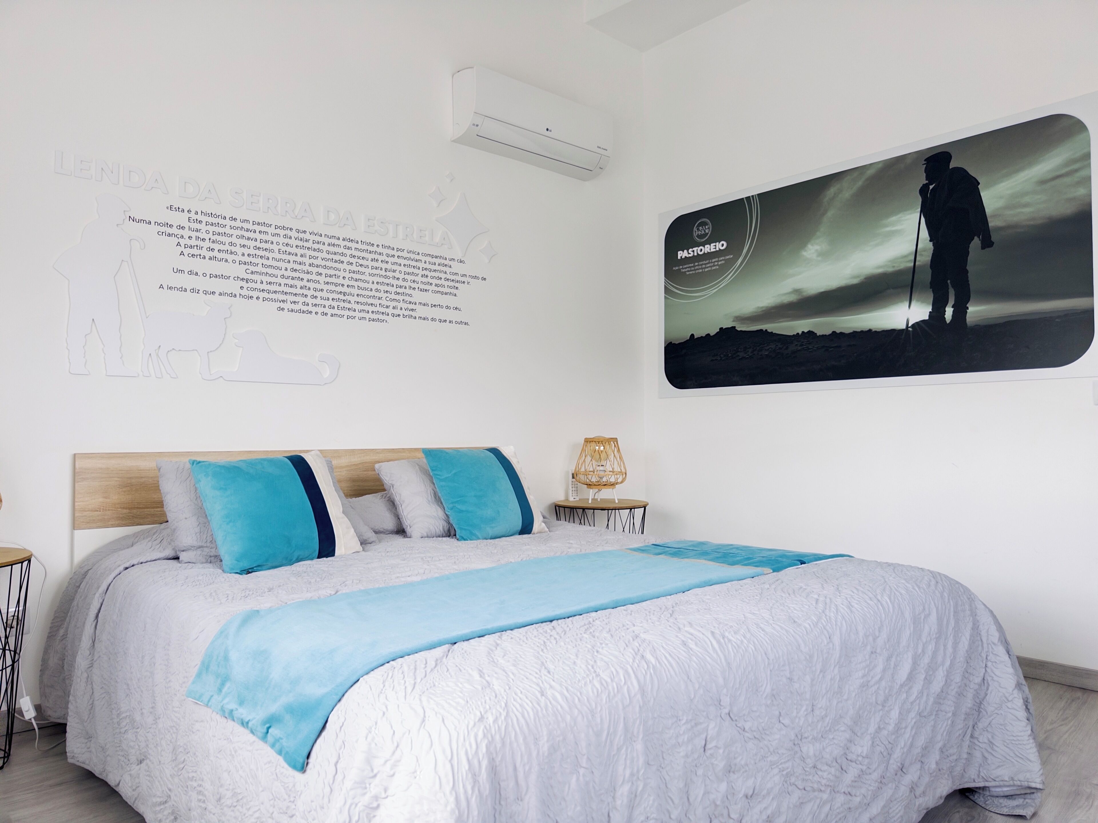 Un dormitorio con una cama, aire acondicionado de pared, un cuadro enmarcado de una persona haciendo senderismo y un cartel de texto en la pared.