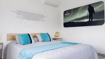 Un dormitorio con una cama, aire acondicionado de pared, un cuadro enmarcado de una persona haciendo senderismo y un cartel de texto en la pared.
