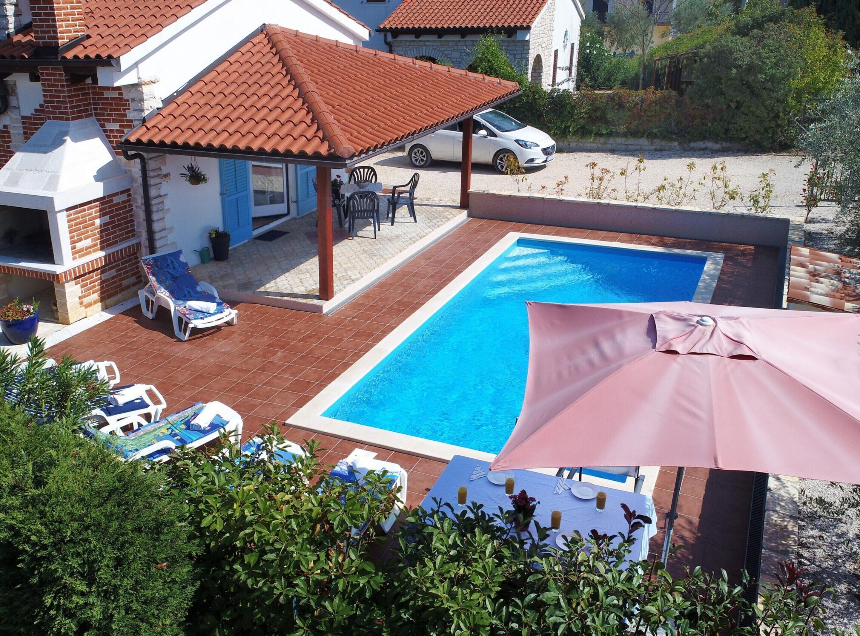 A swimming pool with lounge chairs and umbrellas, a covered patio area, and a parked car.