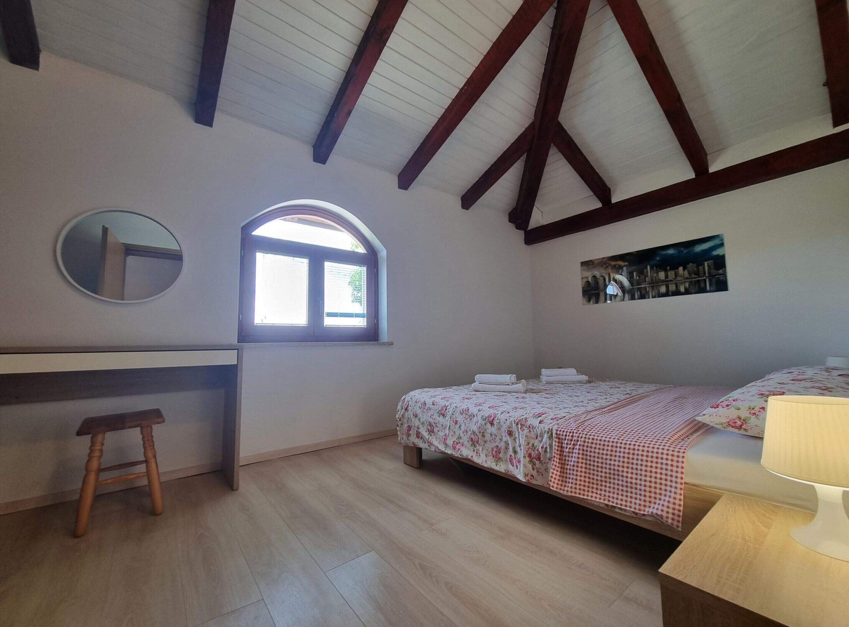 A bedroom with a wooden desk, a small stool, a bed with floral bedding, a window, and a cityscape painting.