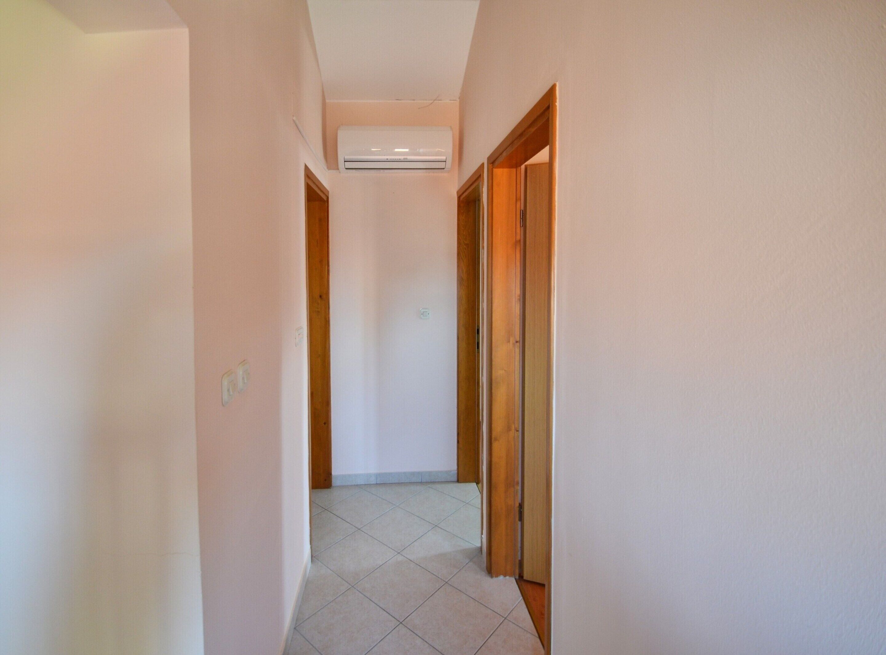 A narrow hallway with a ceiling-mounted air conditioner, wooden doors, and tiled flooring.