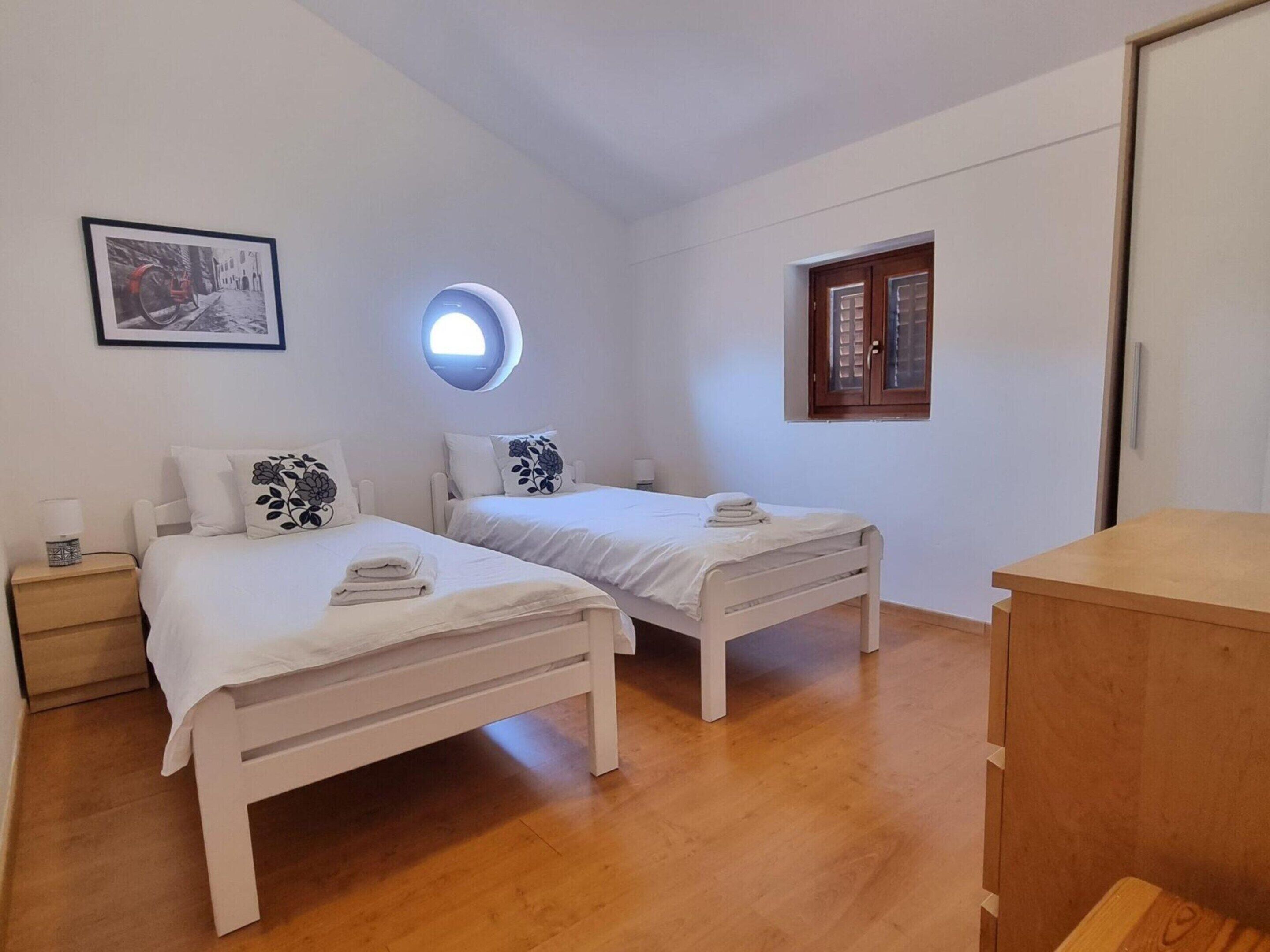 Une chambre avec deux lits, une lucarne ronde, une fenêtre et une commode en bois.