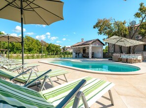 A swimming pool with lounge chairs and umbrellas, a covered seating area, and a stone building in the background. - Villa in Sveti Lovrec Near Ski Resort (Tinjan)