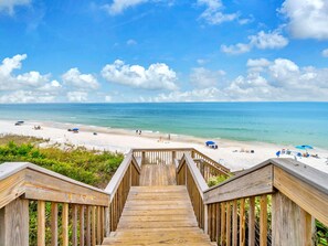 Eine Holztreppe führt zu einem Strand mit blauem Himmel und ruhigem Wasser.