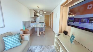 A modern living room with a sofa, a dining table, and a TV mounted on the wall displaying streaming services. - Flat Oceânica Seaside - By Almare (João Pessoa)